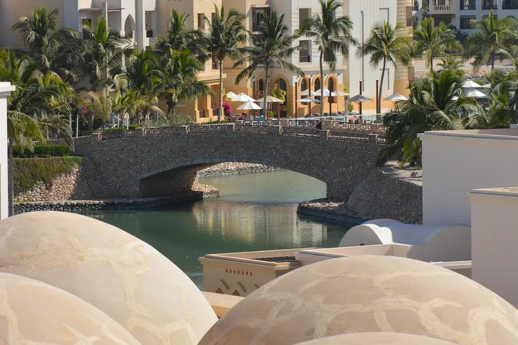 Stone bridge over water at a resort in Muscat, Oman, with palm trees.